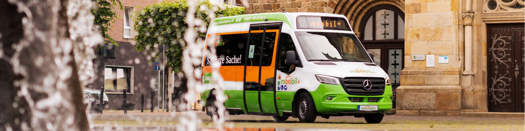 Ein moobil+Bus steht vor einem Gebäude und im Vordergrund ist der Strahl einer Wasserfontäne zu sehen.